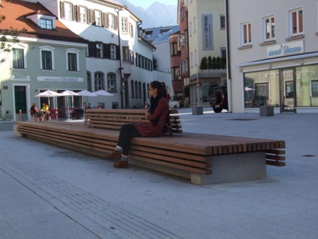 euroform w - Stadtmobiliar - Frau sitzt auf minimalistischer Bank aus Holz und Beton mit indirekter Beleuchtung bei Wiltener Platzl in Innsbruck - Sitzinsel aus Holz und Beton auf öffentlichem Platz in Österreich - customized Stadtmöbel - Sonderlösung für