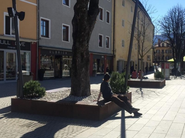 euroform w - Stadtmobiliar - robuste Bank aus hochwertigem Holz für den städtischen Raum mit Planzschale - minimalistischer Sitzgelegenheit aus Holz für draußen - hochwertige Designer Stadtmöbel - Menschen sitzen auf Bank aus Hartholz mit Baum in Mitte