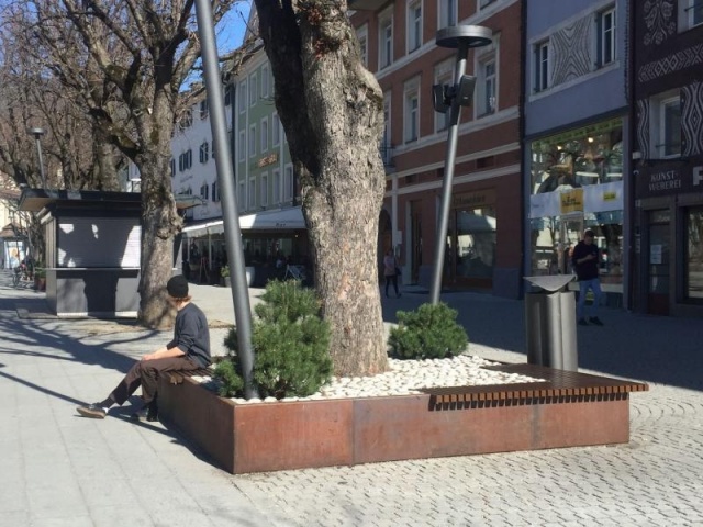 euroform w - Stadtmobiliar - robuste Bank aus hochwertigem Holz für den städtischen Raum mit Planzschale - minimalistischer Sitzgelegenheit aus Holz für draußen - hochwertige Designer Stadtmöbel - Menschen sitzen auf Bank aus Hartholz mit Baum in Mitte