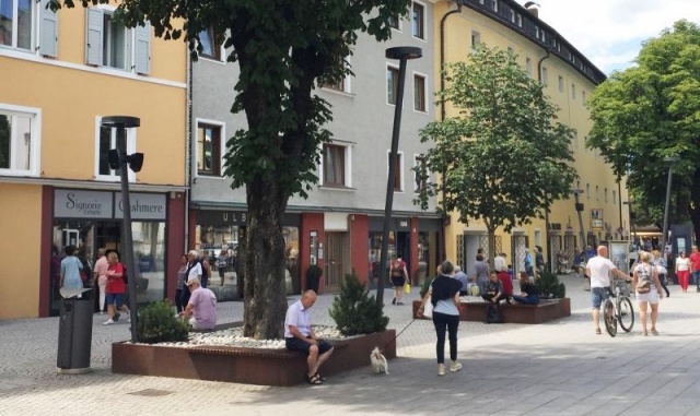 euroform w - Stadtmobiliar - robuste Bank aus hochwertigem Holz für den städtischen Raum mit Planzschale - minimalistischer Sitzgelegenheit aus Holz für draußen - hochwertige Designer Stadtmöbel - Menschen sitzen auf Bank aus Hartholz mit Baum in Mitte