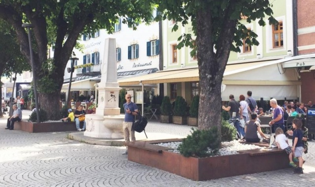 euroform w - street furniture - robust bench made of high quality wood for urban space with planar - minimalist wooden seating for outdoors - high quality designer street furniture - people sit on bench made of hardwood with tree in centre for public park